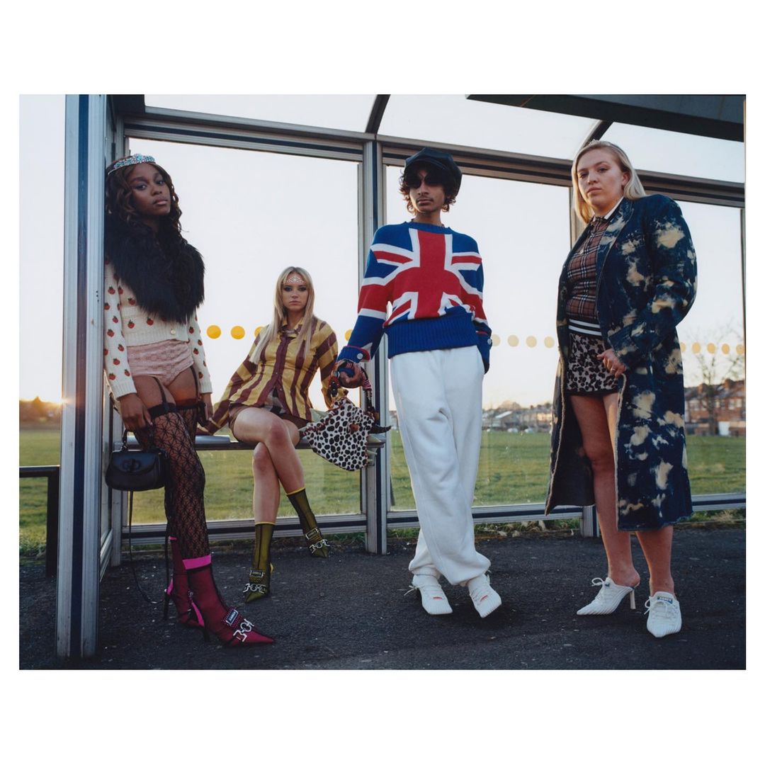 Four stylish people pose at a bus stop wearing knit sweaters, coats, skirts, dresses, and high-heeled shoes in an outdoor scene.