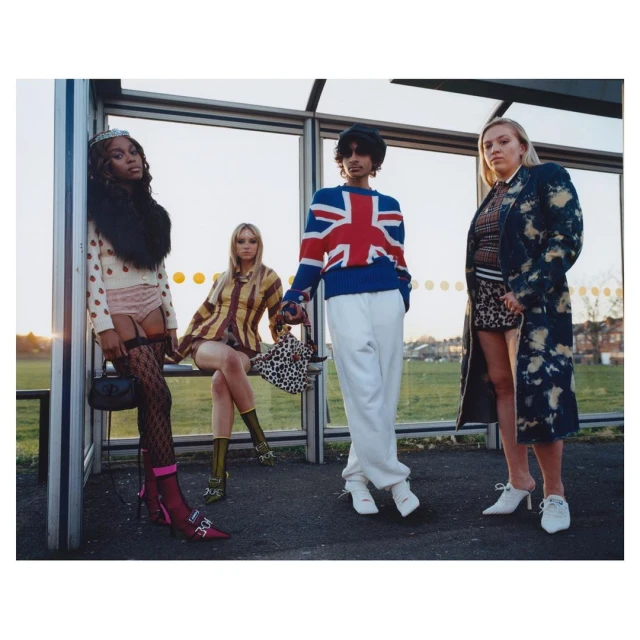 Four stylish people pose at a bus stop wearing knit sweaters, coats, skirts, dresses, and high-heeled shoes in an outdoor scene.