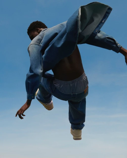 An African American man in denim clothing is angled in the frame against a bright blue sky, with arms and legs outstretched in mid-air.