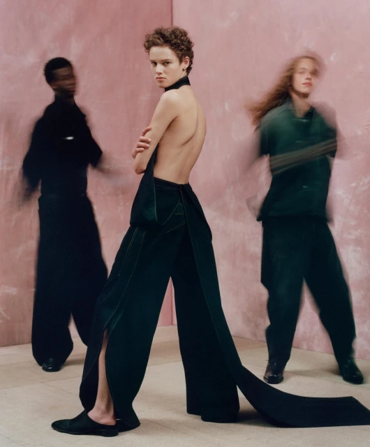 Three fashion models pose in a studio against a pink wall. One model faces the camera, wearing wide-leg pants and a neck wrap.