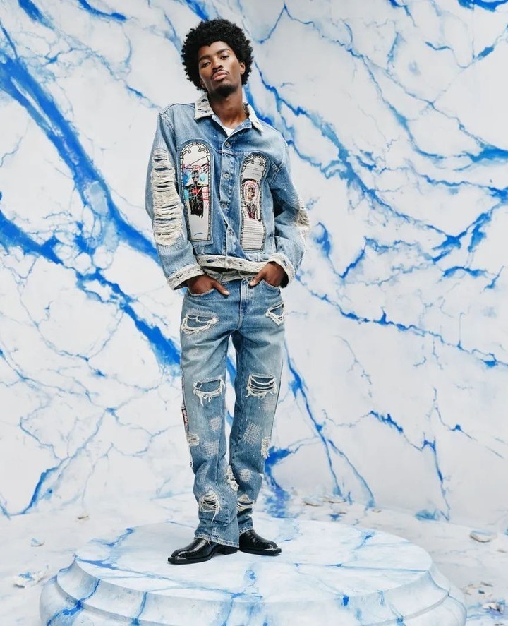 Full-length studio shot of adult male with afro, wearing ripped jeans and denim jacket, hands in pockets, blue marble background.