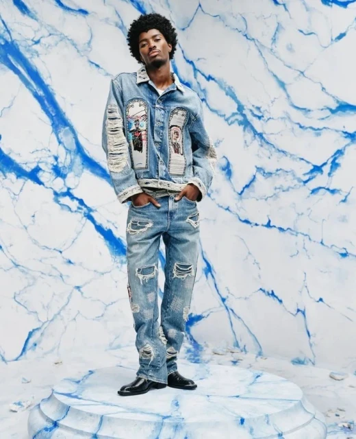 Full-length studio shot of adult male with afro, wearing ripped jeans and denim jacket, hands in pockets, blue marble background.