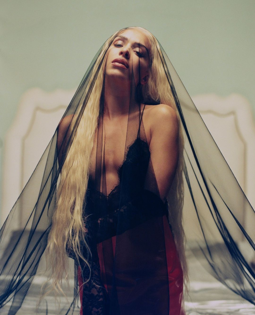 Woman with long blonde hair wearing black lingerie and a veil, posing indoors with a serene expression. Soft, dreamy lighting.