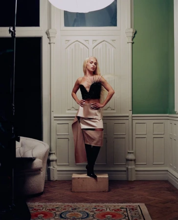 Elegant blonde woman poses in a black top and draped skirt on a wooden box in an ornate studio with high ceilings and rug.