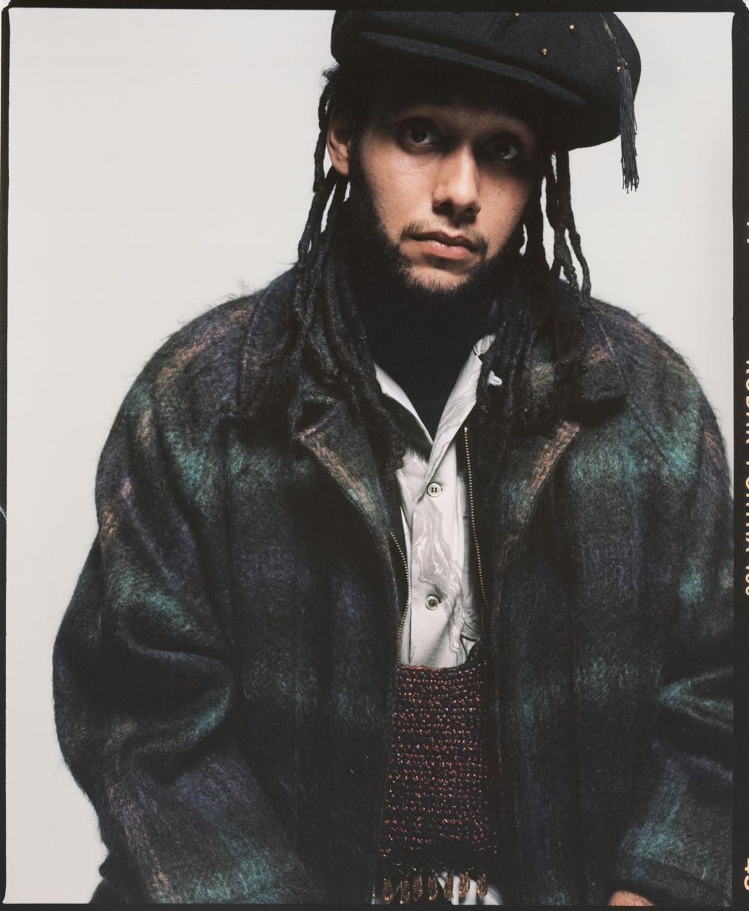 Portrait of a Black adult male in a hat and overcoat with dreadlocks against a plain background in a studio with medium framing.