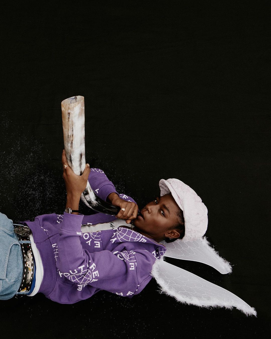 Person in a purple sweatshirt and hat lying on a black surface while holding an animal horn and wearing white angel wings.
