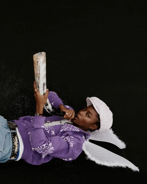 Person in a purple sweatshirt and hat lying on a black surface while holding an animal horn and wearing white angel wings.