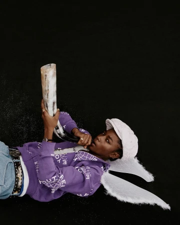 Person in a purple sweatshirt and hat lying on a black surface while holding an animal horn and wearing white angel wings.