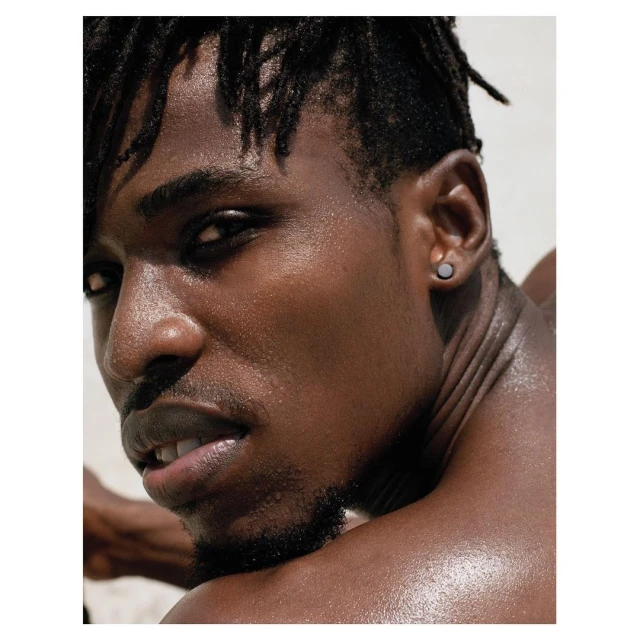 Close-up studio portrait of a young adult Black man with dreadlocks, showcasing his bare shoulders and striking facial features against a neutral backdrop.