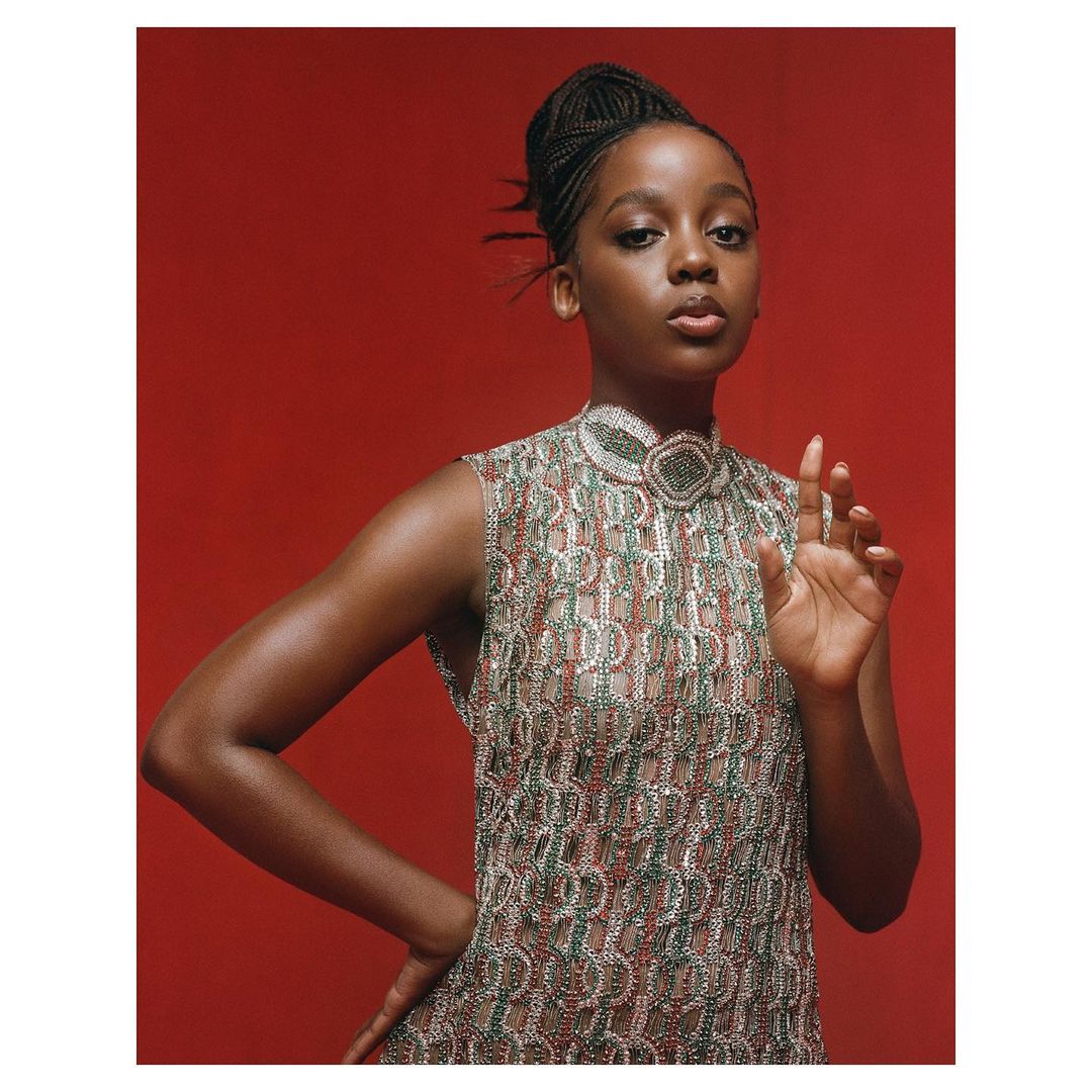A young, dark-skinned woman poses in an embellished sleeveless dress against a solid red backdrop, looking directly at the camera.