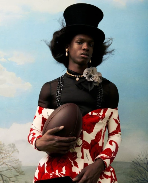 Elegant portrait of a Black adult male holding an American football, wearing a floral dress and black top hat, against a blue sky.