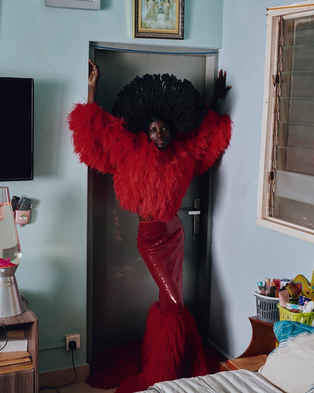 A slender Black model poses in a red feathered top, sequined skirt, and afro hairstyle against a doorway in a home interior.