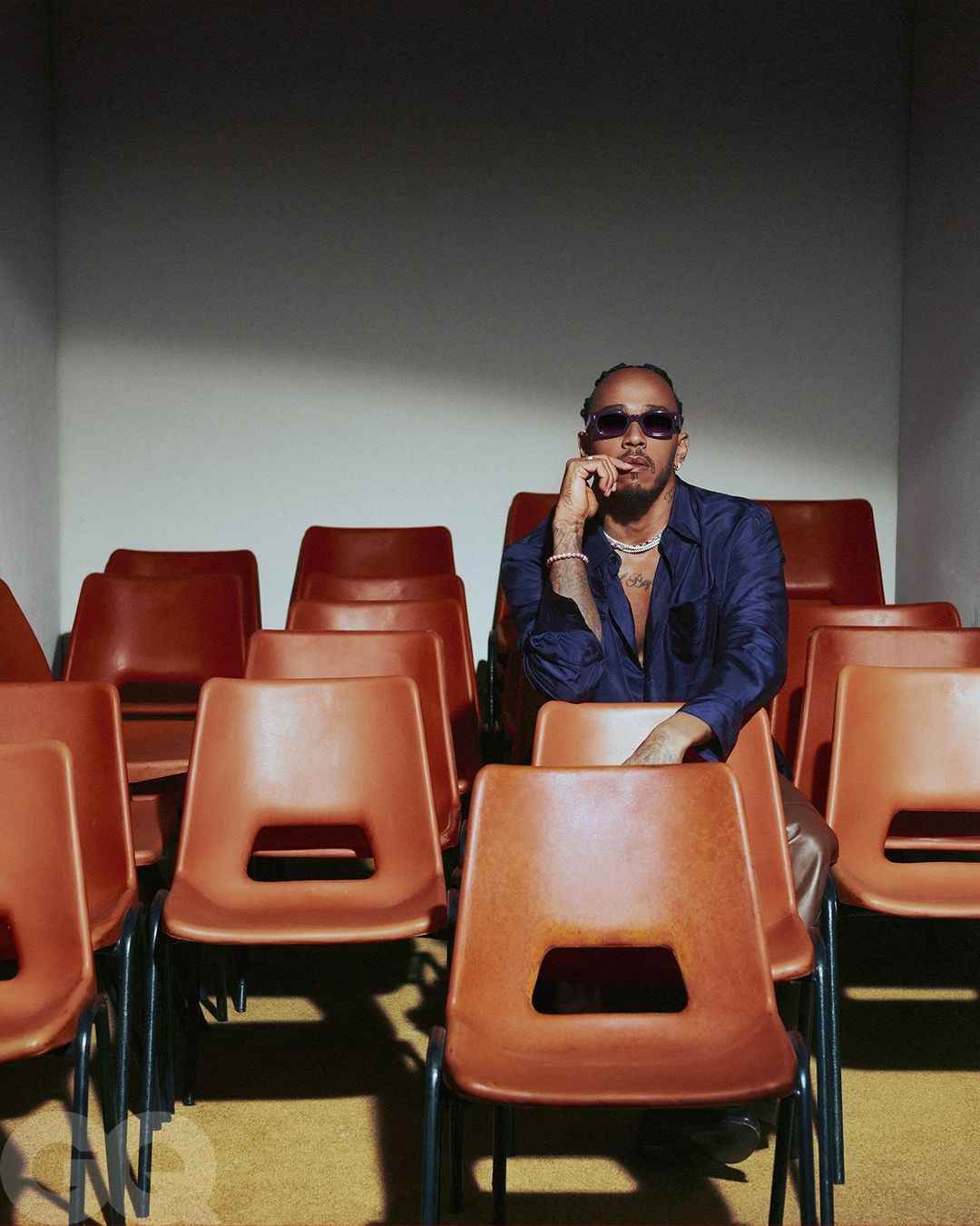 Portrait of adult male, Lewis Hamilton, seated in rows of orange chairs, wearing a navy button-down shirt and tinted sunglasses.