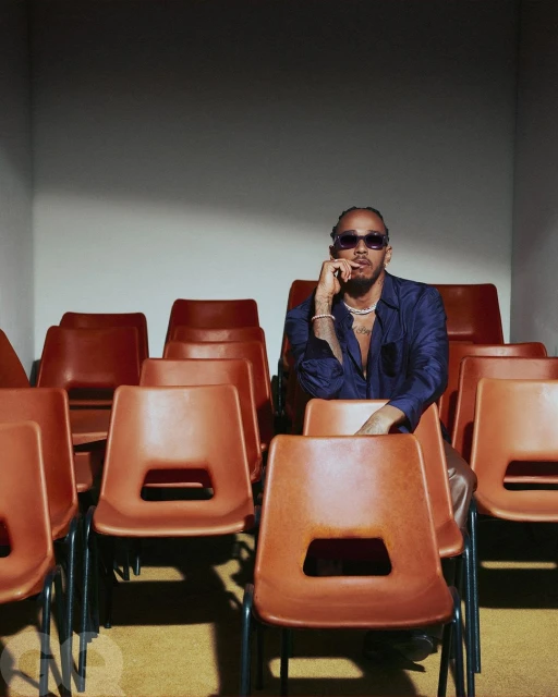 Portrait of adult male, Lewis Hamilton, seated in rows of orange chairs, wearing a navy button-down shirt and tinted sunglasses.