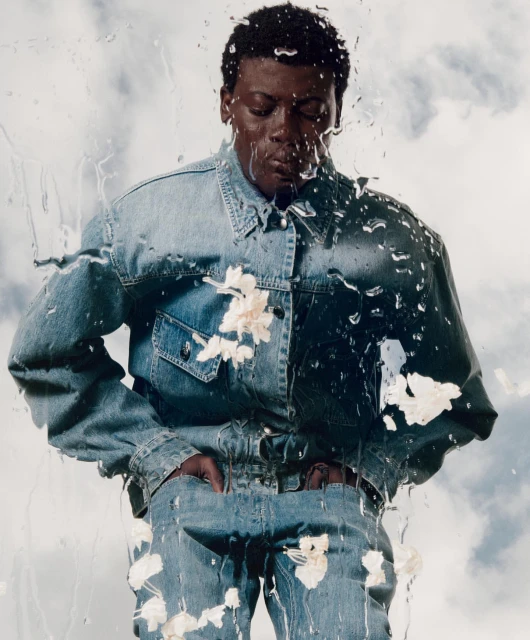 Eye-level portrait of a Black male model in a denim outfit with flowers and water droplets, set against a sky-cloud background.