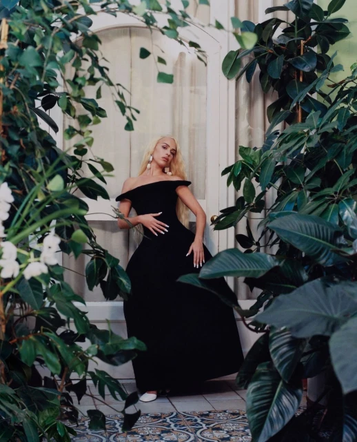 A woman with long blonde hair in a black dress poses in front of an ornate door framed by large green plants and flowers.