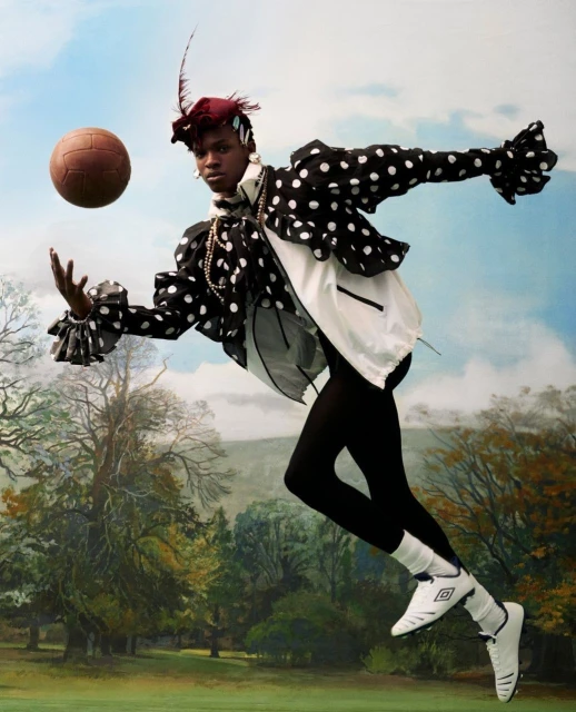 An adult female in elaborate polka dot costume kicks an antique soccer ball in a surreal outdoor setting.