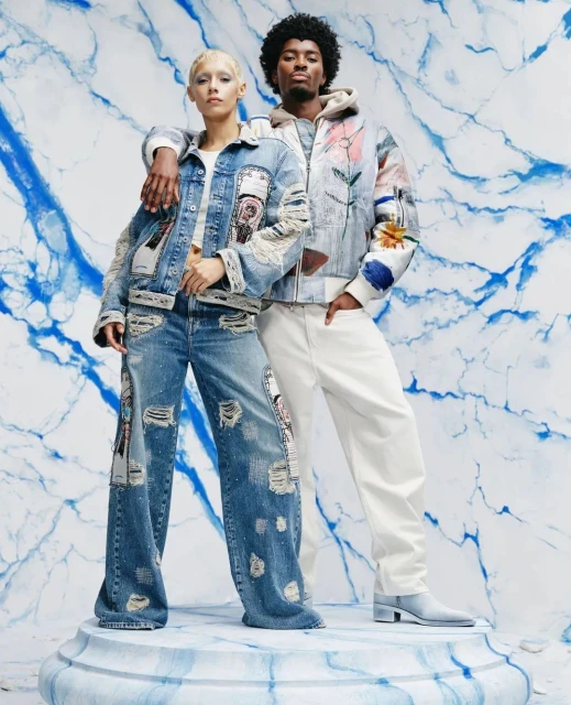 Two fashion models pose in designer denim, a jacket, and white pants against a blue marble backdrop in a studio shot.
