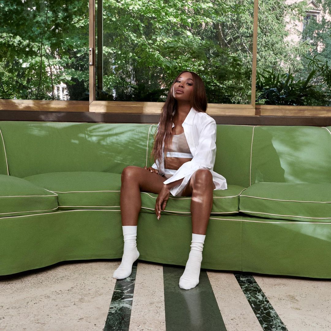 Woman in white shirt sits on a green couch in an indoor setting with a marble floor and a large window overlooking trees.