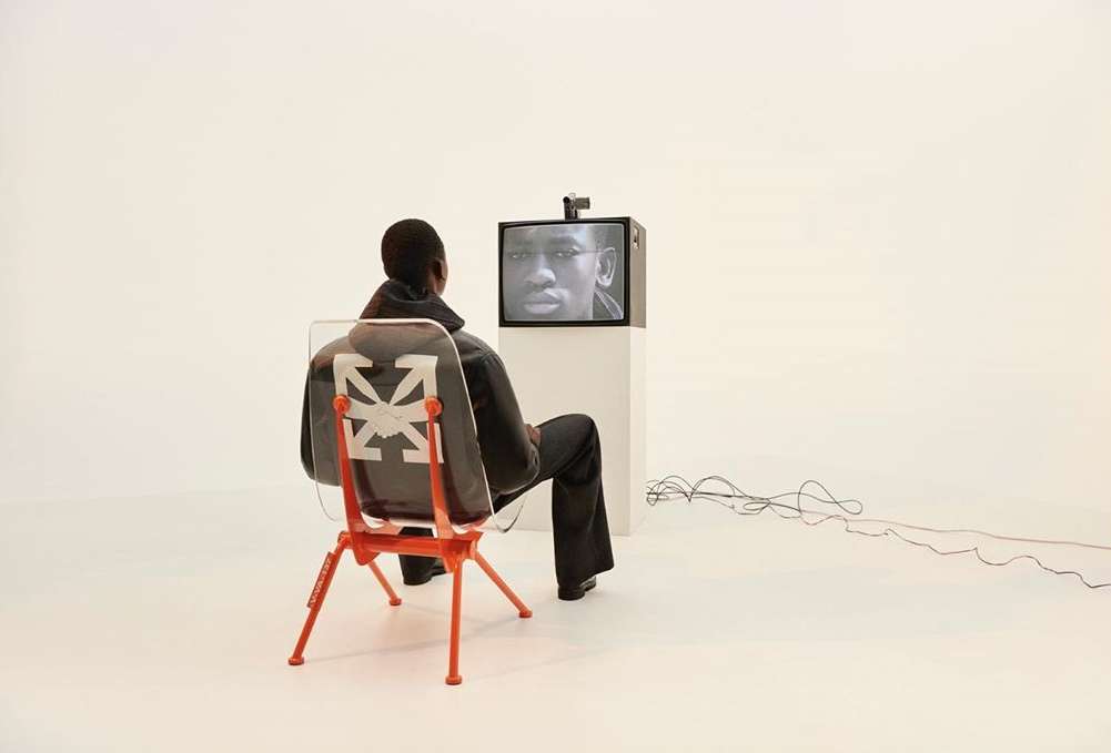 Adult male sits in a chair watching an older television with a person on the screen in a bright white, minimal studio environment.