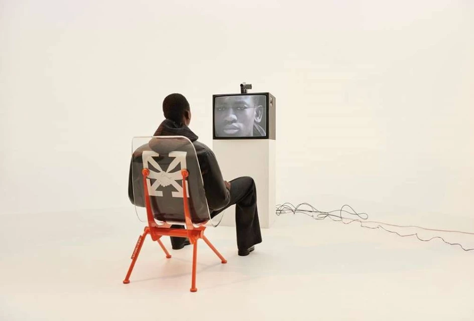 Adult male sits in a chair watching an older television with a person on the screen in a bright white, minimal studio environment.