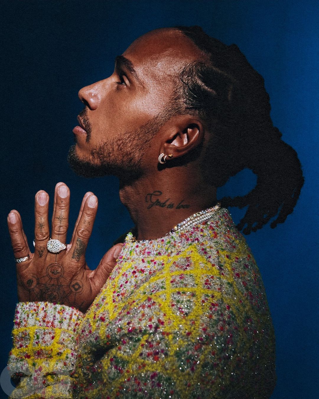 Portrait of a handsome adult male with locs, wearing a colorful knit sweater with hands folded in prayer against a dark blue backdrop.