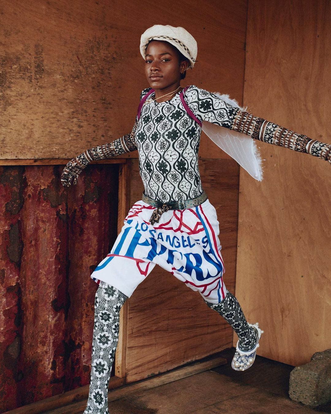 Young adult poses indoors wearing fashion ensemble consisting of patterned top, skirt shorts with text, and matching patterned leggings.