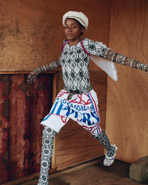 Young adult poses indoors wearing fashion ensemble consisting of patterned top, skirt shorts with text, and matching patterned leggings.