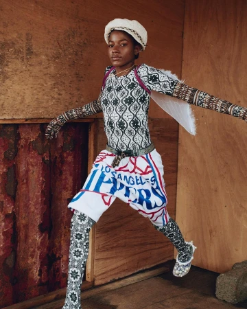 Portrait of a black woman in patterned long-sleeve top and Los Angeles Clippers shorts posing against wooden walls.