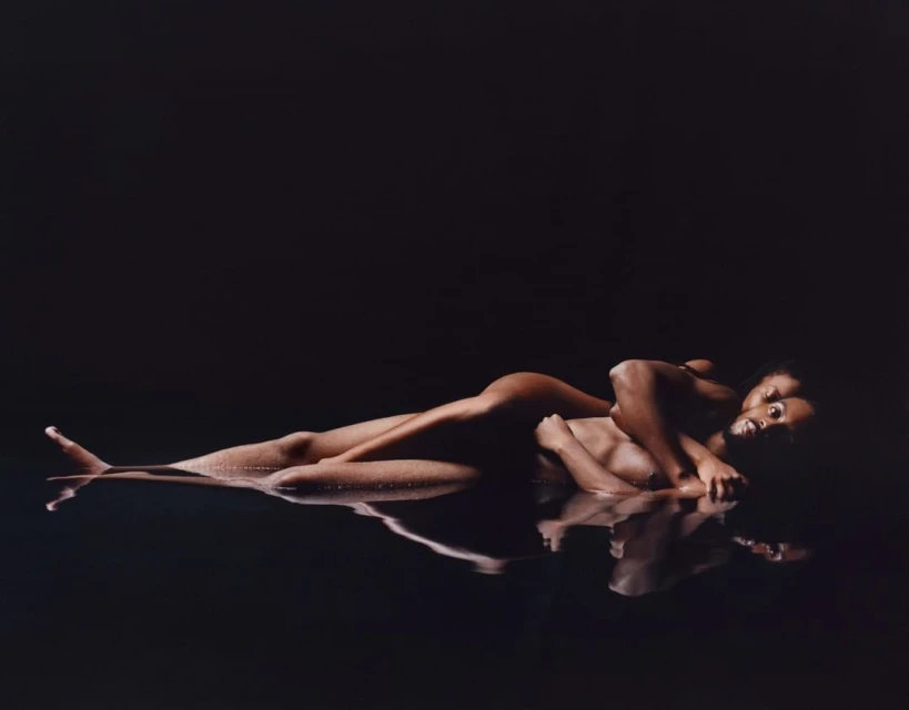 Nude man and woman embrace closely on a reflective surface against a black background, creating a sensual and intimate studio portrait.