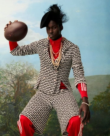 A stylish young Black man poses in a printed suit with a football, set against a painterly sky with clouds and trees, exuding fashion-forward confidence.