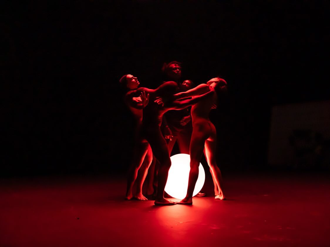 Group of dancers performing with red lighting and a glowing orb in a dark space, highlighting their movements and forms.