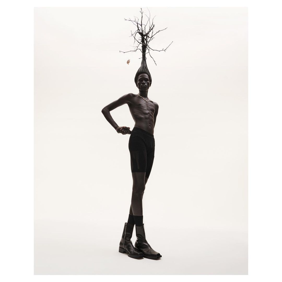Dark-skinned adult male posing with bare torso, wearing dark briefs and boots, under studio lighting, with a headdress of tree branches.