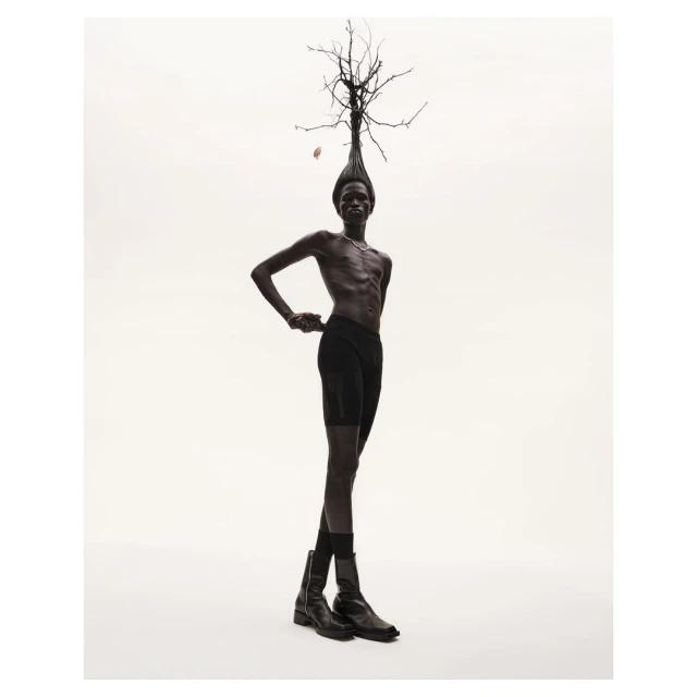 Dark-skinned adult male posing with bare torso, wearing dark briefs and boots, under studio lighting, with a headdress of tree branches.