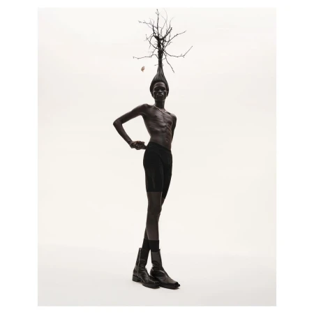 Dark-skinned adult male posing with bare torso, wearing dark briefs and boots, under studio lighting, with a headdress of tree branches.