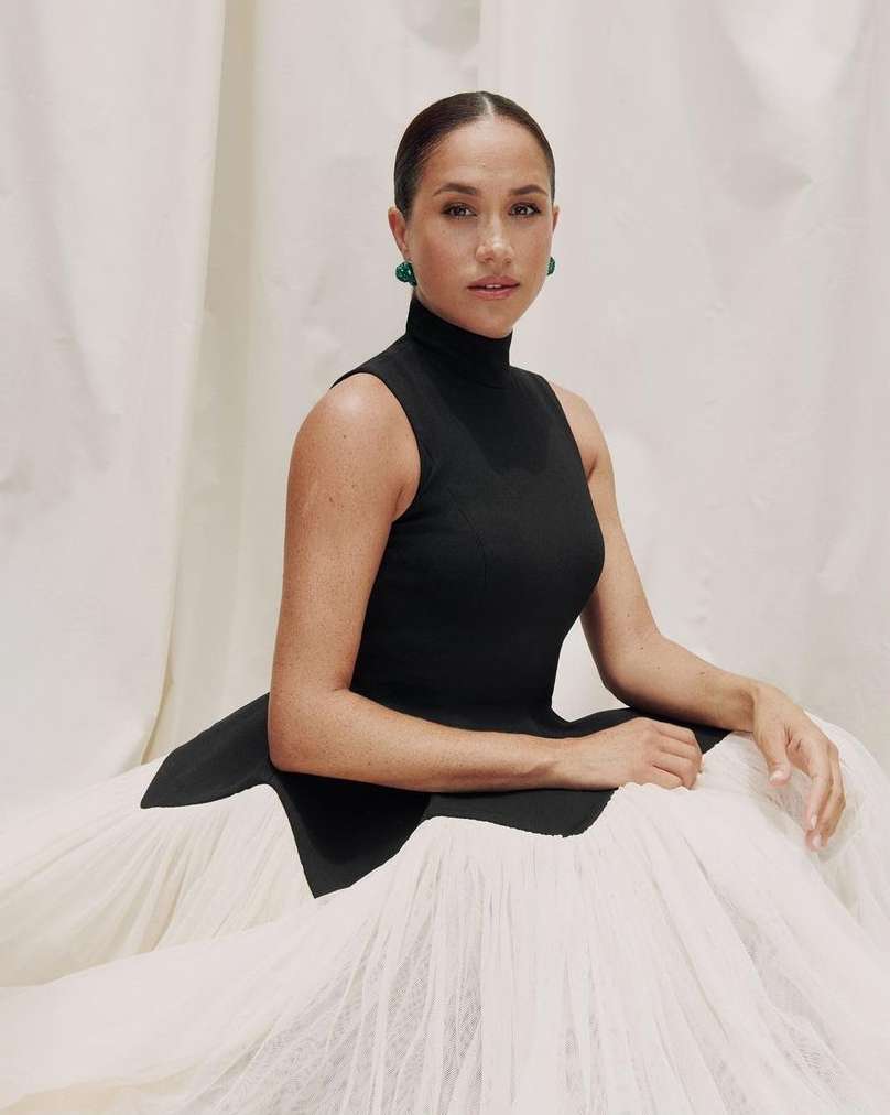 Portrait of a woman with her hair slicked back, wearing a black sleeveless dress and green earrings, posing in front of fabric.