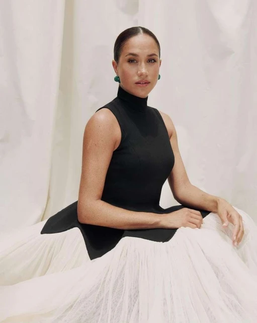 Portrait of a woman with her hair slicked back, wearing a black sleeveless dress and green earrings, posing in front of fabric.