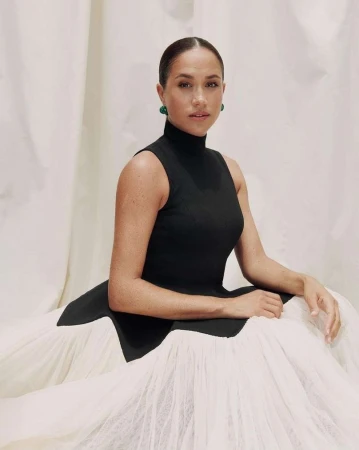 Portrait of a woman with her hair slicked back, wearing a black sleeveless dress and green earrings, posing in front of fabric.