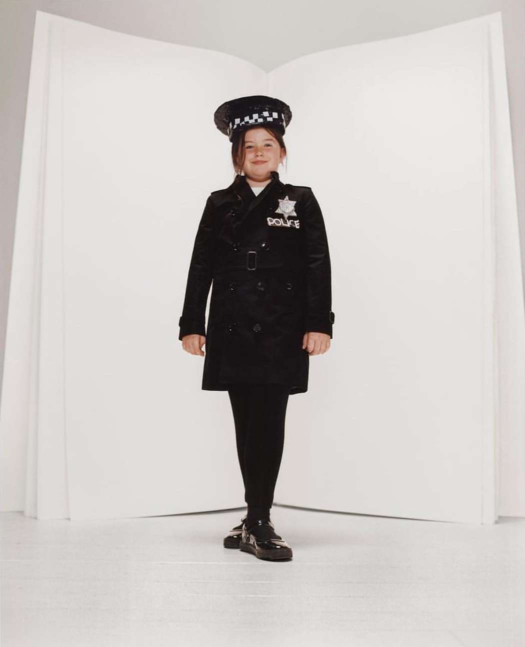 Young girl in a police uniform standing against a white backdrop, posing for a full-length portrait in a studio setting.