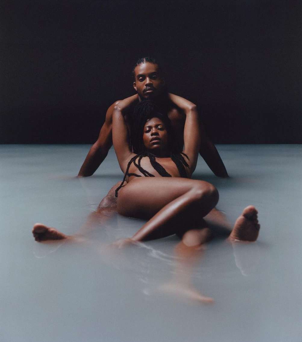 Photo of a shirtless man embracing a woman lying in milky water, looking at the camera with a neutral expression against a dark background.