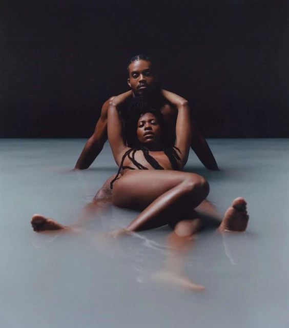 Photo of a shirtless man embracing a woman lying in milky water, looking at the camera with a neutral expression against a dark background.