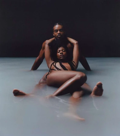 Photo of a shirtless man embracing a woman lying in milky water, looking at the camera with a neutral expression against a dark background.
