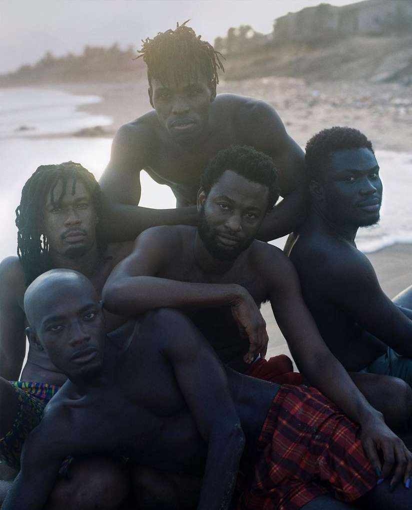 Group of five shirtless men of African descent posing on a sandy beach with ocean waves and an overcast sky in the background.