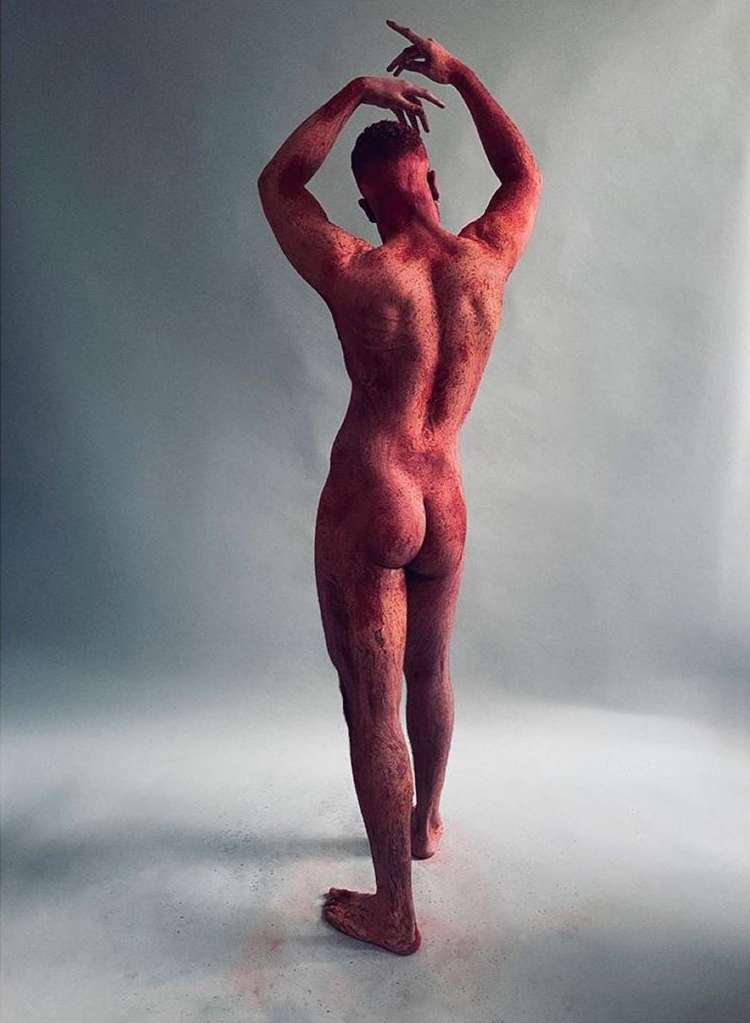 Rear view of a nude adult male figure with red dust, posed with arms overhead in a brightly lit studio setting, monochromatic color grading.