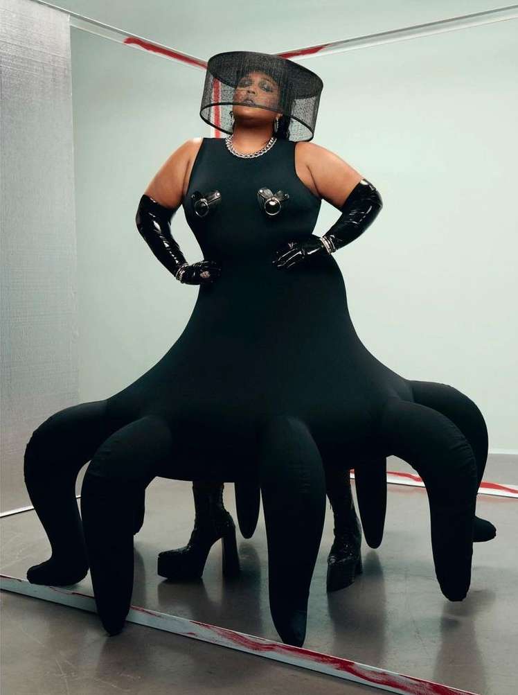Stylish woman poses in a black dress with tentacle-like extensions, complemented by a black hat and gloves in a studio setting.