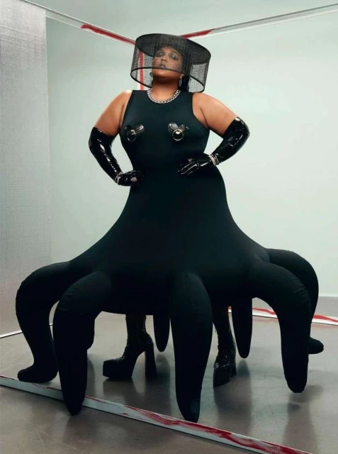 Stylish woman poses in a black dress with tentacle-like extensions, complemented by a black hat and gloves in a studio setting.