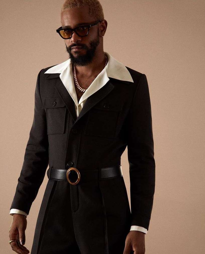 Studio shot of an adult male with blond hair and beard wearing black jumpsuit, yellow sunglasses, and gold chain against beige backdrop.
