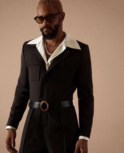 Studio shot of an adult male with blond hair and beard wearing black jumpsuit, yellow sunglasses, and gold chain against beige backdrop.