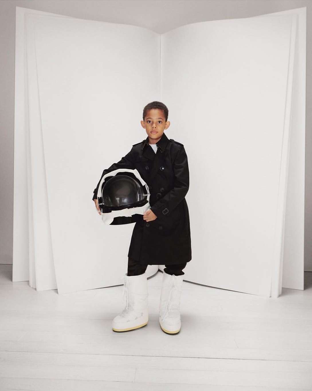 Young boy in a black trench coat holds a space helmet and wears white moon boots in a bright studio with a blank white background.