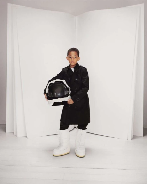 Young boy in a black trench coat holds a space helmet and wears white moon boots in a bright studio with a blank white background.
